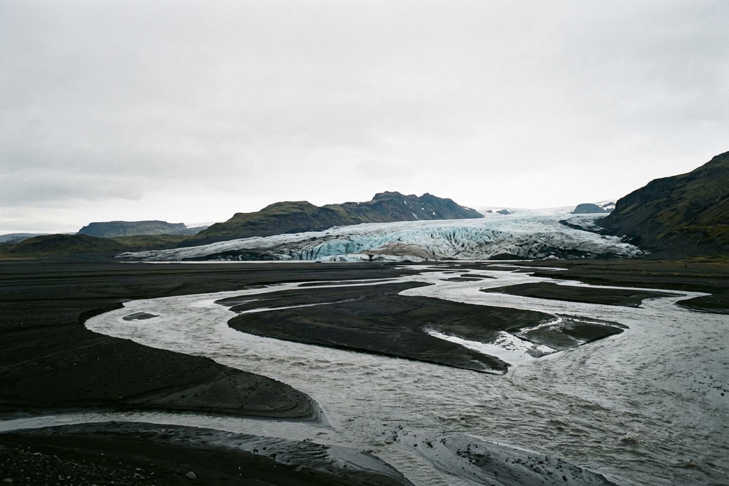 A glacier tongue reaching down to a black sand plain braided with meltwater channels